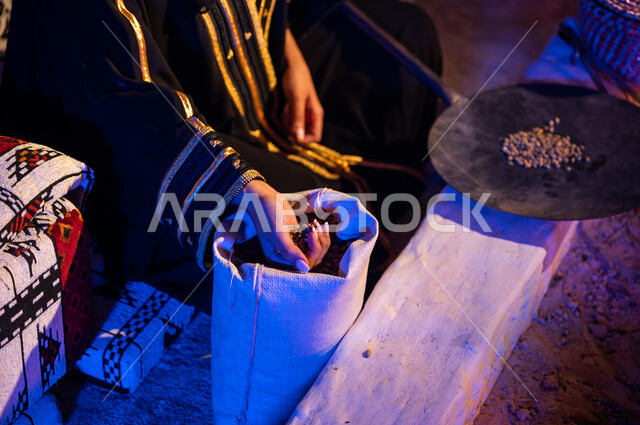 أخذ البن من الكيس ووضعه في المحماس لتحميصه على شعلة النار في موقد طيني، صورة مقربة لإمرأه عربيه خليجية سعودية  تستخدم المعاميل لإعداد القهوة بالطريقة التقليدية على الحطب داخل خيمة شعبيه تراثية، منتج محلي ومحاصيل زراعية وطنية، أسلوب الحياة القديمه في الباديه