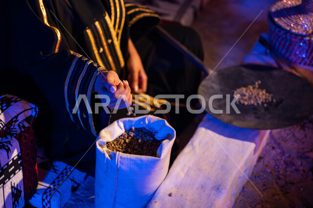 أخذ البن من الكيس ووضعه في المحماس لتحميصه على شعلة النار في موقد طيني، صورة مقربة لإمرأه عربيه خليجية سعودية  تستخدم المعاميل لإعداد القهوة بالطريقة التقليدية على الحطب داخل خيمة شعبيه تراثية، منتج محلي ومحاصيل زراعية وطنية، أسلوب الحياة القديمه في الباديه
