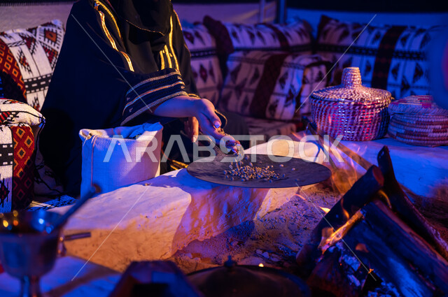 Using ma'amil to prepare coffee in the traditional way on firewood inside a traditional folk tent. A close-up picture of a Saudi Arabian Gulf woman placing coffee beans in ma'amil to roast them over a fire in a clay stove. A local product and national agricultural crops. The ancient way of life in the desert.