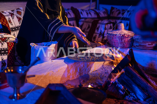 وضع حبوب البن في المحماس لتحميصها على شعلة النار في موقد طيني، صورة مقربة لإمرأه عربيه خليجية سعودية ترتدي العباءة تستخدم المعاميل لإعداد القهوة بالطريقة التقليدية على الحطب داخل خيمة شعبيه تراثية، منتج محلي ومحاصيل زراعية وطنية، أسلوب الحياة القديمه في الباديه