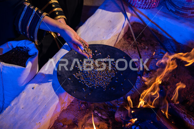 Using ma'amil to prepare coffee in the traditional way on firewood inside a traditional folk tent. A close-up picture of a Saudi Arabian Gulf woman placing coffee beans in ma'amil to roast them over a fire in a clay stove. A local product and national agricultural crops. The ancient way of life in the desert.
