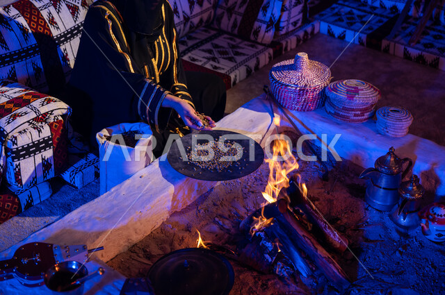 وضع حبوب البن في المحماس لتحميصها على شعلة النار في موقد طيني، صورة مقربة لإمرأه عربيه خليجية سعودية ترتدي العباءة تستخدم المعاميل لإعداد القهوة بالطريقة التقليدية على الحطب داخل خيمة شعبيه تراثية، منتج محلي ومحاصيل زراعية وطنية، أسلوب الحياة القديمه في الباديه
