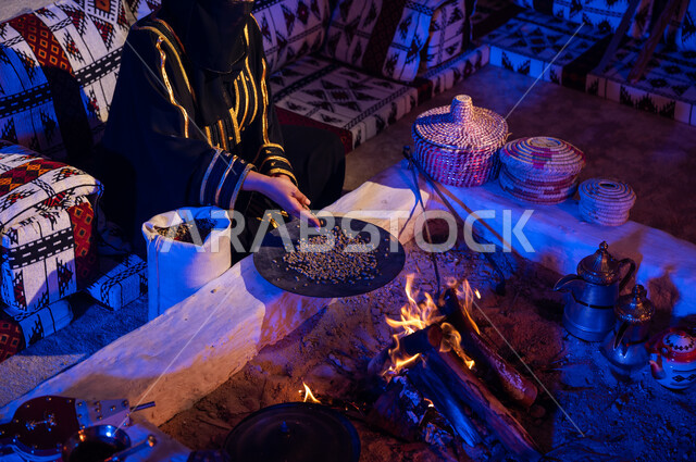 Putting the coffee beans in the roaster to roast them over the flame of a clay stove. A close-up photo of a Saudi Arabian Gulf woman wearing an abaya using ma’ameel to prepare coffee in the traditional way on firewood inside a popular heritage tent. A local product and national agricultural crops. The ancient way of life in the desert.
