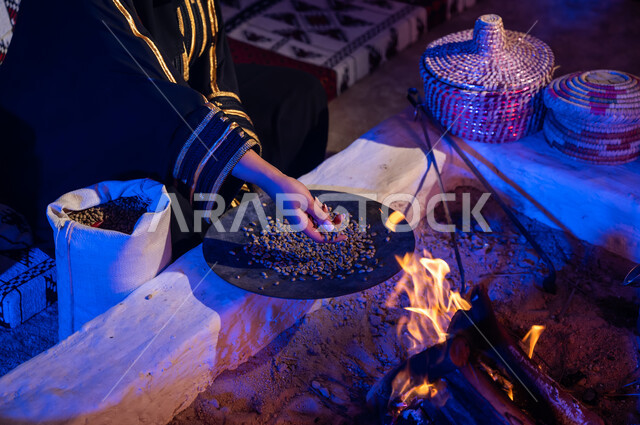 وضع حبوب البن في المحماس لتحميصها على شعلة النار في موقد طيني، صورة مقربة لإمرأه عربيه خليجية سعودية ترتدي العباءة تستخدم المعاميل لإعداد القهوة بالطريقة التقليدية على الحطب داخل خيمة شعبيه تراثية، منتج محلي ومحاصيل زراعية وطنية، أسلوب الحياة القديمه في الباديه