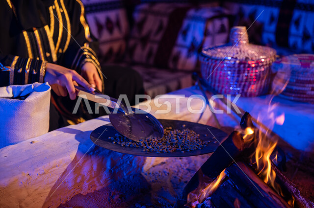 منتج محلي ومحاصيل زراعية وطنية، استخدام المعاميل لتحضير القهوة بالطريقة التقليدية على الحطب في موقد طيني داخل خيمة شعبيه تراثية، أسلوب الحياة القديمه في الباديه، صورة مقربة لأيدي إمرأة عربية خليجيه سعودية تقوم بتحميص حبوب البن في المحماس على شعلة النار