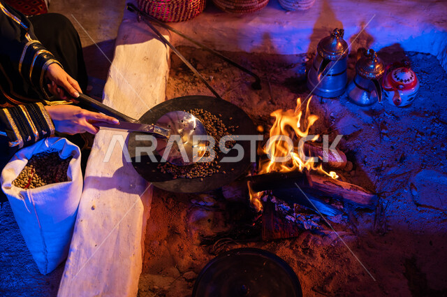 استعمال المعاميل لتحضير القهوة بالطريقة التقليدية على الحطب في موقد طيني داخل خيمة شعبيه تراثية، أسلوب الحياة القديمه في الباديه، صورة مقربة لأيدي إمرأة عربية خليجيه سعودية تقوم بتحميص حبوب البن في المحماس على شعلة النار، منتج محلي ومحاصيل زراعية وطنية