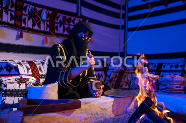 استخدام المعاميل لإعداد القهوة بالطريقة التقليدية على الحطب في موقد طيني داخل خيمة شعبيه تراثية، إمرأه عربيه خليجية سعودية ترتدي العباءه السوداء والبرقع تقوم بطحن حبوب البن في المهباج النحاسي، منتج محلي ومحاصيل زراعية وطنية، اسلوب الحياة القديمه في الباديه،