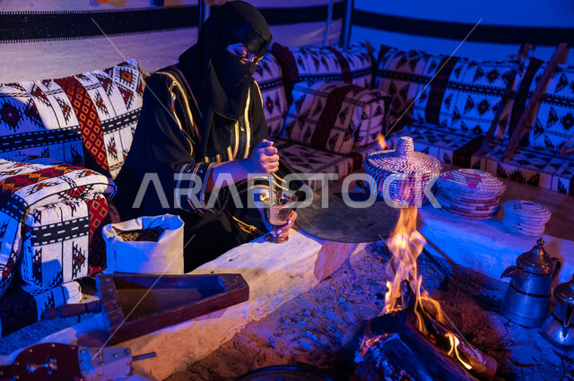استخدام المعاميل لإعداد القهوة بالطريقة التقليدية على الحطب في موقد طيني داخل خيمة شعبيه تراثية، إمرأه عربيه خليجية سعودية ترتدي العباءه السوداء والبرقع تقوم بطحن حبوب البن في المهباج النحاسي، منتج محلي ومحاصيل زراعية وطنية، اسلوب الحياة القديمه في الباديه،