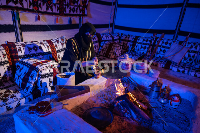 استخدام المعاميل لإعداد القهوة بالطريقة التقليدية على الحطب في موقد طيني داخل خيمة شعبيه تراثية، إمرأه عربيه خليجية سعودية ترتدي العباءه السوداء والبرقع تقوم بطحن حبوب البن في المهباج النحاسي، منتج محلي ومحاصيل زراعية وطنية، اسلوب الحياة القديمه في الباديه،