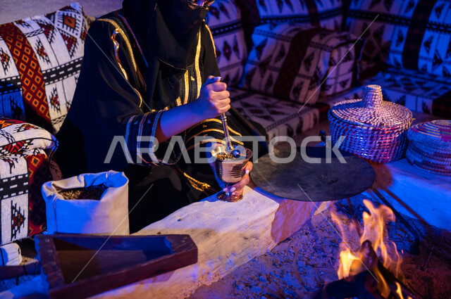 نمط الحياة القديمه في الباديه، طحن حبوب البن في النجر النحاسي، صورة مقربة لإمرأه عربيه خليجية سعودية ترتدي العباءه السوداء تقوم باستخدام المعاميل لإعداد القهوة بالطريقة التقليدية على الحطب في موقد طيني داخل خيمة شعبيه تراثية، منتج محلي ومحاصيل زراعية وطنية