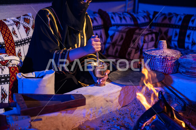 The ancient way of life in the desert, grinding coffee beans in a copper pot, a close-up picture of a Saudi Gulf Arab woman wearing a black abaya, using ma’ameel to prepare coffee in the traditional way on firewood in a clay stove inside a traditional popular tent, a local product and national agricultural crops.