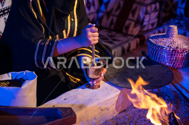 أسلوب الحياة القديمه في الباديه، طحن حبوب البن في النجر النحاسي، صورة مقربة لإمرأه عربيه خليجية سعودية ترتدي العباءه السوداء تقوم باستخدام المعاميل لإعداد القهوة بالطريقة التقليدية على الحطب في موقد طيني داخل خيمة شعبيه تراثية، منتج محلي ومحاصيل زراعية وطنية