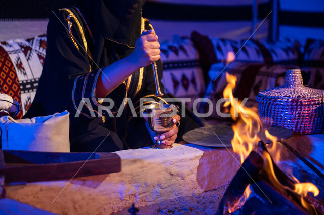 طحن حبوب البن في النجر النحاسي، صورة مقربة لإمرأه عربيه خليجية سعودية ترتدي العباءه والبرقع تقوم باستخدام المعاميل لإعداد القهوة بالطريقة التقليدية على الحطب في موقد طيني داخل خيمة شعبيه تراثية، منتج محلي ومحاصيل زراعية وطنية، أسلوب الحياة القديمه في الباديه