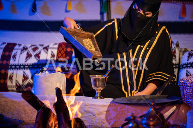 توارث مهارة صنع القهوة العربية ، مشروبات وطنية تراثية شعبية, استعمال المعاميل لصنع القهوة بالطريقة التقليدية على الحطب في موقد طيني، صورة مقربة لامرأة عربية خليجية سعودية ترتدي العباءة والبرقع تقوم بنقل حبوب البن من المبرد الى النجر لطحنها