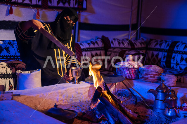 الاستمتاع بصنع القهوة العربية داخل خيمة تراثية، استعمال المعاميل لصنع القهوة بالطريقة التقليدية على الحطب في موقد طيني، صورة مقربة لامرأة عربية خليجية سعودية ترتدي العباءة والبرقع تقوم بنقل حبوب البن من المبرد الى النجر لطحنها، محاصيل زراعية وطنية محلية