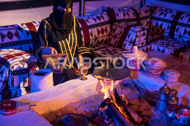 التقاليد السعودية الأصيلة في صنع القهوة العربية، استعمال المعاميل لصنع القهوة بالطريقة التقليدية على الحطب في موقد طيني، امرأة عربية خليجية سعودية ترتدي العباءة والبرقع تقوم بنقل حبوب البن من المبرد الى النجر لطحنها، محاصيل زراعية وطنية محلية
