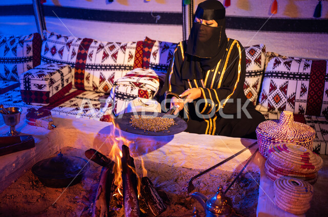 تحضير القهوة بالطريقة التقليدية على الحطب في موقد طيني باستخدام المعاميل، أسلوب الحياة القديمة في البادية، إمرأة عربية خليجية سعودية ترتدي العباءة السوداء والبرقع تقوم بتحميص حبوب البن في المحماس على شعلة النار داخل خيمة تراثية، منتج محلي ومحاصيل زراعية وطنية