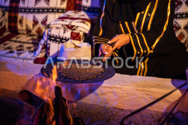 Using ma'ameel to prepare coffee in the traditional way on firewood in a clay stove, the ancient way of life in the desert, a close-up of a Saudi Arabian Gulf woman wearing an abaya, roasting coffee beans in mahamas over a fire inside a heritage tent, a local product and national agricultural crops.