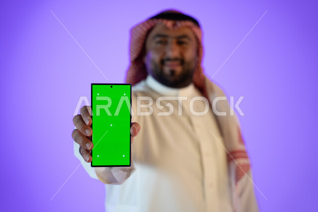 مفهوم التسويق الالكتروني, التسوق والشراء عن بعد, بورتريه لرجل أعمال عربي سعودي خليجي مبتسم يستخدام هاتفه المحمول بخلفية خضراء, كروما، استخدام تقنية السمارت على الجوال, خلفية ملونة