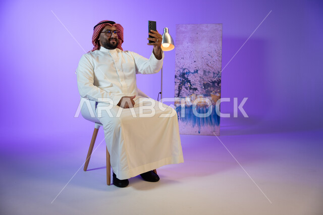 التقاط صور سيلفي تذكارية, مفهوم صلة الرحم و التواصل مع الأصدقاء والأقارب بإيماءات السرور, رجل عربي خليجي سعودي يرتدي الثوب والشماغ الأحمر يقوم بإجراء مكالمة فيديو عبر الجوال, دردشات وحوارات ودية مع الأصحاب, الجلوس واستخدام تطبيقات التواصل الاجتماعي