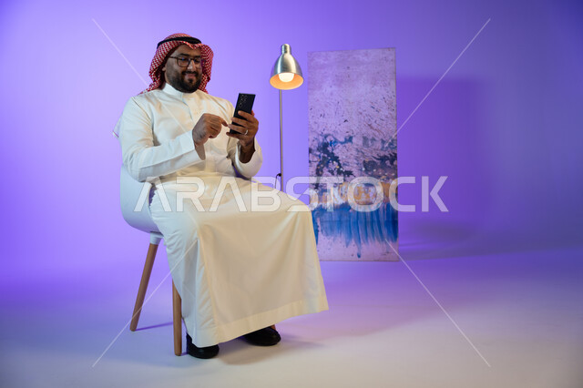التسوق الإلكتروني السريع والآمن, إرسال رسالة نصية عبر الجوال, رجل عربي خليجي سعودي مبتسم يرتدي الثوب والشماغ التقليدي يجلس على الكرسي و يقوم بمتابعة أخبار السوشال ميديا عبر الهاتف المحمول, مفهوم صلة الرحم و التواصل مع الأصدقاء والأقارب