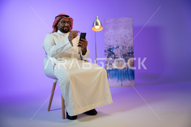 التسوق الإلكتروني السريع والآمن, إرسال رسالة نصية عبر الجوال, رجل عربي خليجي سعودي مبتسم يرتدي الثوب والشماغ التقليدي يجلس على الكرسي و يقوم بمتابعة أخبار السوشال ميديا عبر الهاتف المحمول, مفهوم صلة الرحم و التواصل مع الأصدقاء والأقارب