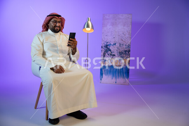 إجراء مكالمة فيديو عبر الجوال, رجل عربي خليجي سعودي مبتسم يرتدي الثوب والشماغ التقليدي يجلس على الكرسي و يقوم بمتابعة أخبار السوشال ميديا عبر الهاتف المحمول, مفهوم صلة الرحم و التواصل مع الأصدقاء والأقارب, التسوق الإلكتروني السريع والآمن