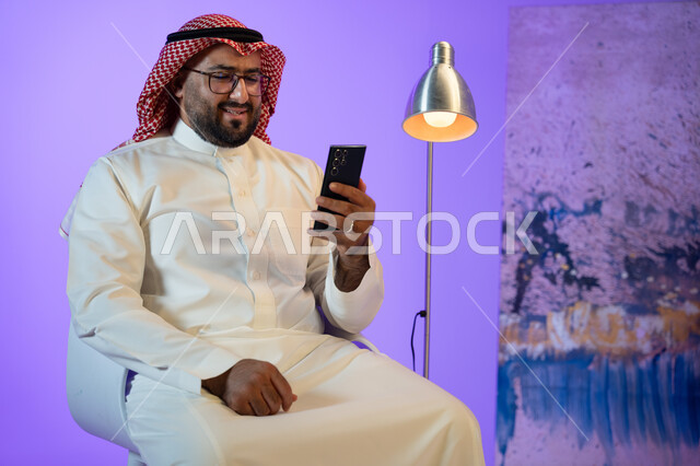 إجراء مكالمة فيديو عبر الجوال, رجل عربي خليجي سعودي مبتسم يرتدي الثوب والشماغ التقليدي يجلس على الكرسي و يقوم بمتابعة أخبار السوشال ميديا عبر الهاتف المحمول, مفهوم صلة الرحم و التواصل مع الأصدقاء والأقارب, التسوق الإلكتروني السريع والآمن