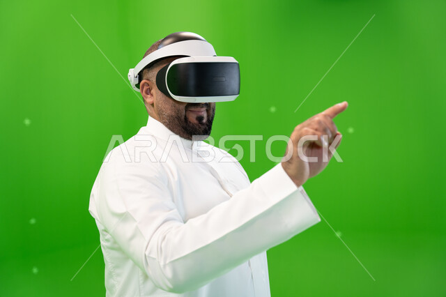 إيماءات التفاعل مع الواقع الافتراضي, التسلية والترفيه بالتقنيات الحديثة, الضغط على شاشة هولوجرامية بإصبع السبابة، بورتريه مقرب من الجانب لرجل عربي خليجي سعودي يرتدي الزي التقليدي و نظارة الواقع الإفتراضي ثلاثية الأبعاد، خلفية خضراء، كروما
