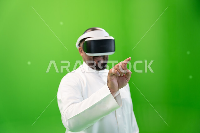 التسلية والترفيه بالتقنيات الحديثة, إيماءات التفاعل مع الواقع الافتراضي, الضغط على شاشة هولوجرامية بإصبع السبابة، بورتريه مقرب  لرجل عربي خليجي سعودي يرتدي الزي التقليدي و نظارة الواقع الإفتراضي ثلاثية الأبعاد، خلفية خضراء، كروما