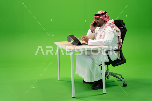 الرد على الاتصالات الهاتفية, العمل في بيئة مريحة, استخدام التقنيات الحديثة لتسهيل الأعمال, بورتريه من الجانب لموظف عربي سعودي خليجي يرتدي الثوب التقليدي يجلس في مكتبه ويتحدث عبر الجوال, مهن ووظائف سعودية مكتبية, خلفية خضراء, كروما.