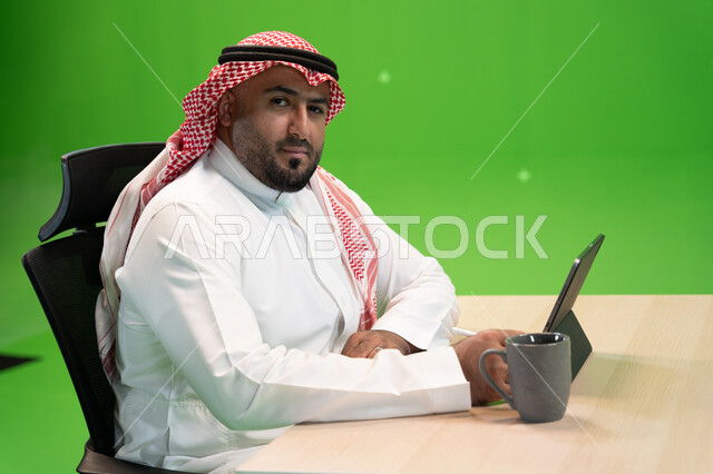 الاستمتاع بالعمل في بيئة مريحة, استخدام الأجهزة والتقنيات الحديثة لتسهيل الأعمال, بورتريه من الجانب لموظف عربي سعودي خليجي يرتدي الثوب التقليدي السعودي يجلس في مكتبه ويستخدم التابلت, مهن ووظائف سعودية مكتبية, النظر الى الكاميرا , خلفية خضراء, كروما.