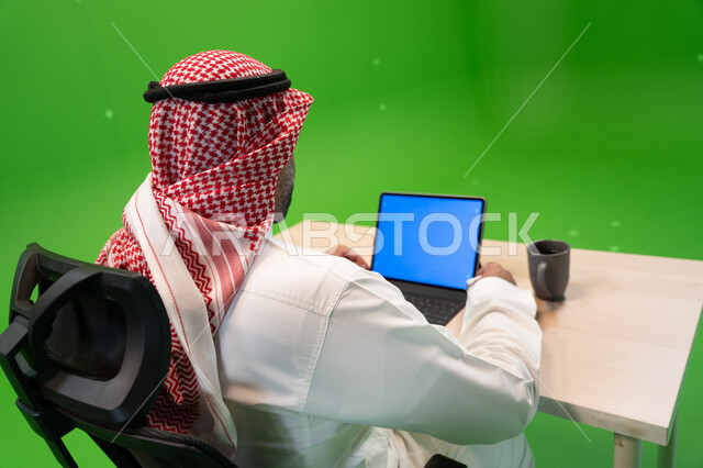 استخدام الأجهزة والتقنيات الحديثة لتسهيل الأعمال, إجراء مقابلات عمل أونلاين, بورتريه من الخلف لموظف عربي سعودي خليجي يرتدي الثوب التقليدي السعودي يجلس في مكتبه ويستخدم التابلت بشاشة زرقاء فارغة, مهن ووظائف سعودية مكتبية, خلفية خضراء, كروما.