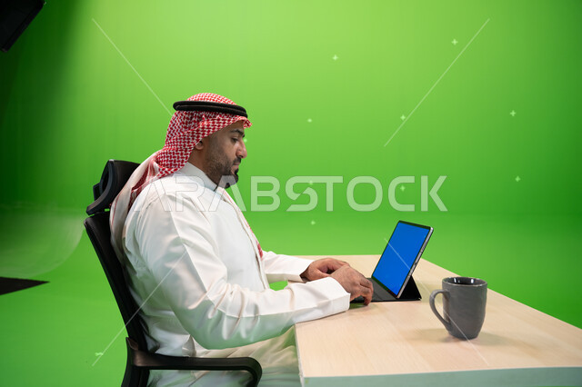 إجراء مقابلات عمل أونلاين, استخدام الأجهزة والتقنيات الحديثة لتسهيل الأعمال, بورتريه من الجانب لموظف عربي سعودي خليجي يرتدي الثوب التقليدي السعودي يجلس في مكتبه ويستخدم التابلت بشاشة زرقاء فارغة, مهن ووظائف سعودية مكتبية, خلفية خضراء, كروما.