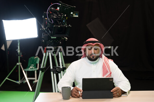 Reading the news summary using a tablet, behind the scenes presenting programs and news bulletins, a Saudi Gulf Arab broadcaster and media personality wearing a shemagh and traditional thobe presenting a television bulletin in front of the camera, world n