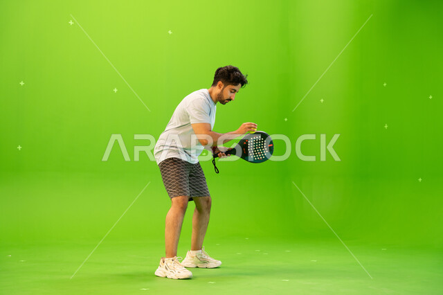 إيماءات تدل على الحماس والاندماج بالتمرين، رياضات شبابية وأنشطة ترفيهية، التدرب على إتقان لعبة البادل، إجراء تمرينات رياضة المضرب، لاعب بادل عربي خليجي سعودي يرتدي ملابس رياضية و يلعب بالكرة والمضرب ، صورة الجسم بالطول الكامل, خلفية خضراء, كروما