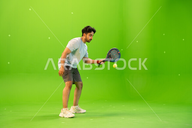 التدرب على إتقان لعبة البادل، رياضات شبابية وأنشطة ترفيهية، إجراء تمرينات رياضة المضرب، لاعب بادل عربي خليجي سعودي يرتدي ملابس رياضية و يلعب بالكرة والمضرب ، إيماءات تدل على الحماس والاندماج بالتمرين، صورة الجسم بالطول الكامل, خلفية خضراء, كروما