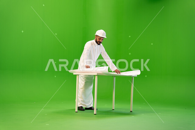 Auditing architectural plans and designs, following up on engineering projects in the Kingdom, a side portrait of a Saudi Arabian Gulf engineer wearing a traditional dress and a protective helmet and standing in front of the table supervising the work sit