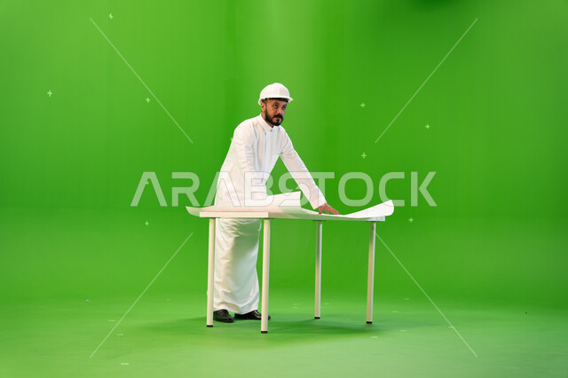 Evaluating and managing architectural designs, following up on engineering projects in the Kingdom, a side portrait of a Saudi Gulf Arab engineer wearing a traditional dress and a protective helmet and standing in front of the table supervising the work s