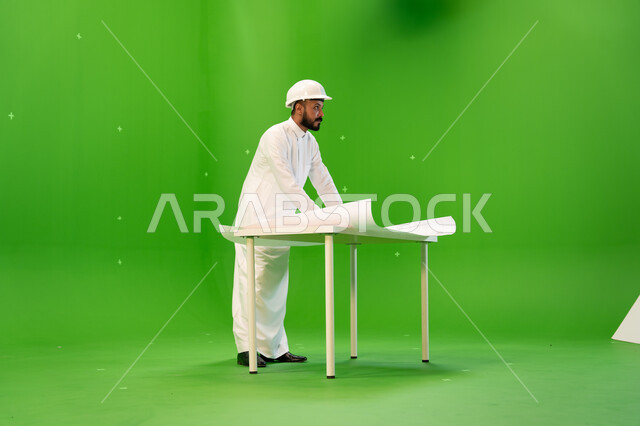 تقييم وإدارة التصاميم المعمارية, متابعة المشاريع الهندسية في المملكة، بورتريه من الجانب لمهندس عربي خليجي سعودي يرتدي الثوب التقليدي و خوذة الحماية ويقف أمام الطاولة يقوم بالإشراف على موقع العمل، صورة الجسم بالطول الكامل, خلفية خضراء، كروما.