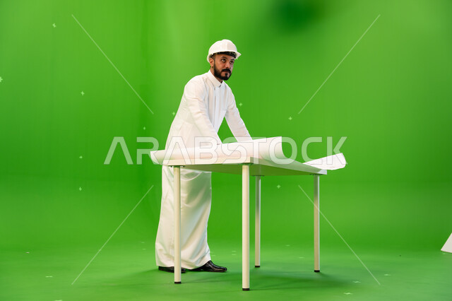 متابعة المشاريع الهندسية في المملكة، تدقيق المخططات والتصاميم المعمارية, بورتريه من الجانب لمهندس عربي خليجي سعودي يرتدي الثوب التقليدي و خوذة الحماية ويقف أمام الطاولة يقوم بالإشراف على موقع العمل، صورة الجسم بالطول الكامل, خلفية خضراء، كروما.