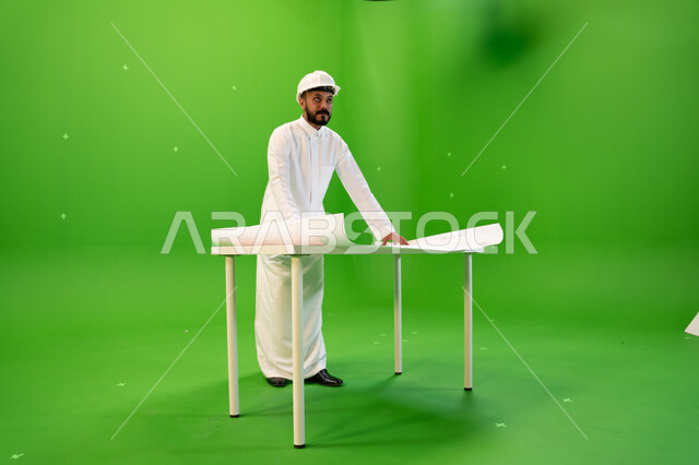 متابعة المشاريع الهندسية في المملكة، تدقيق المخططات والتصاميم المعمارية, بورتريه من الجانب لمهندس عربي خليجي سعودي يرتدي الثوب التقليدي و خوذة الحماية ويقف أمام الطاولة يقوم بالإشراف على موقع العمل، صورة الجسم بالطول الكامل, خلفية خضراء، كروما.