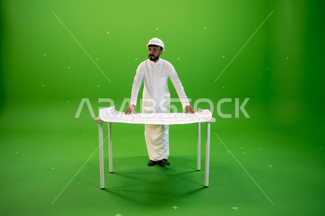 تدقيق المخططات والتصاميم المعمارية, متابعة المشاريع الهندسية في المملكة، بورتريه  لمهندس عربي خليجي سعودي يرتدي الثوب التقليدي و خوذة الحماية ويقف أمام الطاولة يقوم بالإشراف على موقع العمل، صورة الجسم بالطول الكامل, خلفية خضراء، كروما.