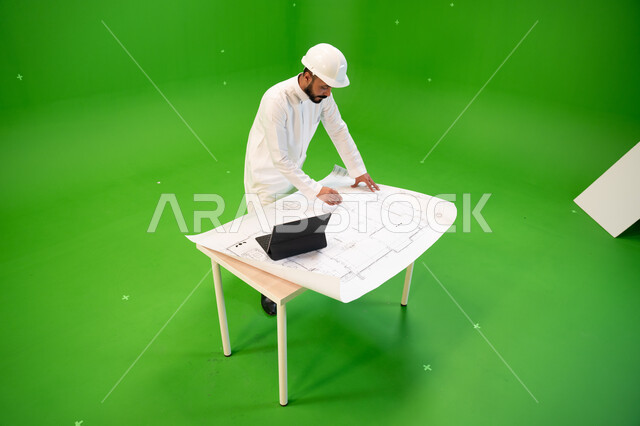 Managing engineering projects using a tablet, a portrait from above of a Saudi Arabian Gulf engineer wearing a traditional dress and a protective helmet and standing at the work site, following up on the progress of construction projects, Saudi profession