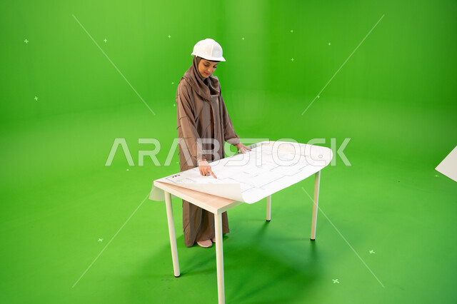 الإشراف على المخططات و التصاميم المعمارية, متابعة المشاريع الهندسية في المملكة، بورتريه من الجانب لمهندسة عربية خليجية سعودية ترتدي العباءة و خوذة الحماية وتقف أمام الطاولة في موقع العمل، صورة الجسم بالطول الكامل, خلفية خضراء، كروما.