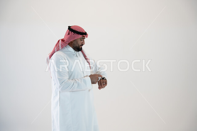 تيسير وتسهيل المهام اليومية باستخدام أحدث التقنيات, تتبع الإشعارات عبر الساعات الحديثة, بورتريه من الجانب لرجل عربي خليجي سعودي يرتدي الشماغ والزي التقليدي ويلبس في يده ساعة ذكية  ويقوم بالنقر عليها, خلفية بيضاء