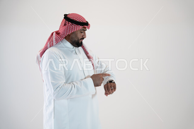 إيماءات النقر والتفاعل مع الساعة الذكية, تيسير وتسهيل المهام اليومية باستخدام أحدث التقنيات, تتبع الإشعارات عبر الساعات الحديثة, بورتريه من الجانب لرجل عربي خليجي سعودي يرتدي الشماغ والثوب التقليدي ويلبس في يده ساعة ذكية, خلفية بيضاء