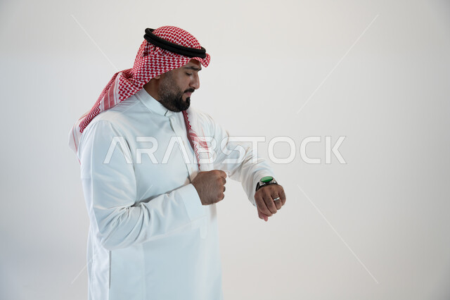 Facilitating and facilitating daily tasks using the latest technologies, tracking notifications through modern watches, a portrait from the side of a Saudi Gulf Arab man wearing a shemagh and traditional dress and wearing a smart watch in his hand and clicking on it, white background
