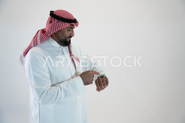 Clicking gestures and interacting with the smart watch, facilitating and facilitating daily tasks using the latest technologies, tracking notifications through modern watches, portrait from the side of a Saudi Gulf Arab man wearing a shemagh and traditional thobe and a smart watch in his hand, white background