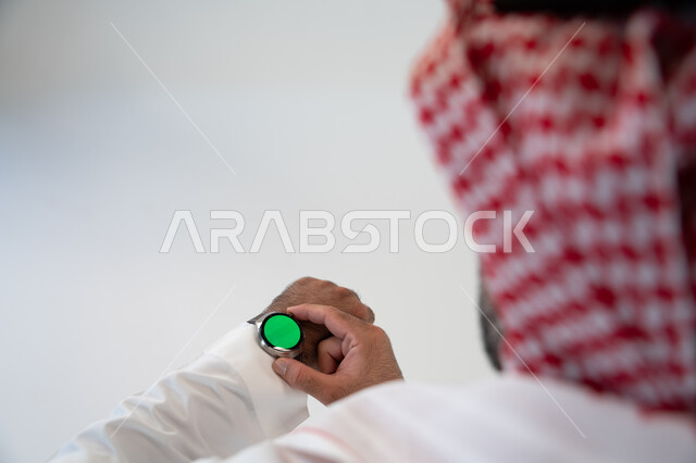 تسهيل إنجاز الأمور الشخصية باستخدام أحدث التقنيات, تتبع الإشعارات عبر الساعات الحديثة, بورتريه مقرب من الخلف لرجل عربي خليجي سعودي يرتدي الشماغ والزي التقليدي ويلبس في يده ساعة ذكية بشاشة خضراء كروما ويقوم بضبطها,  خلفية بيضاء